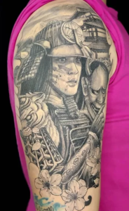 tatuagem oriental em preto e cinza de samurai segurando máscara hannya, ao fundo um templo e flores sakura