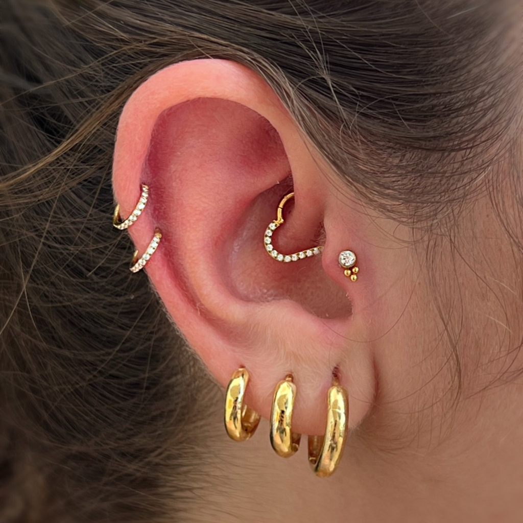 orelha com dois piercings de argola dourados com zircônias, piercing de coração no daith, tragus e brincos de argola no lóbulo